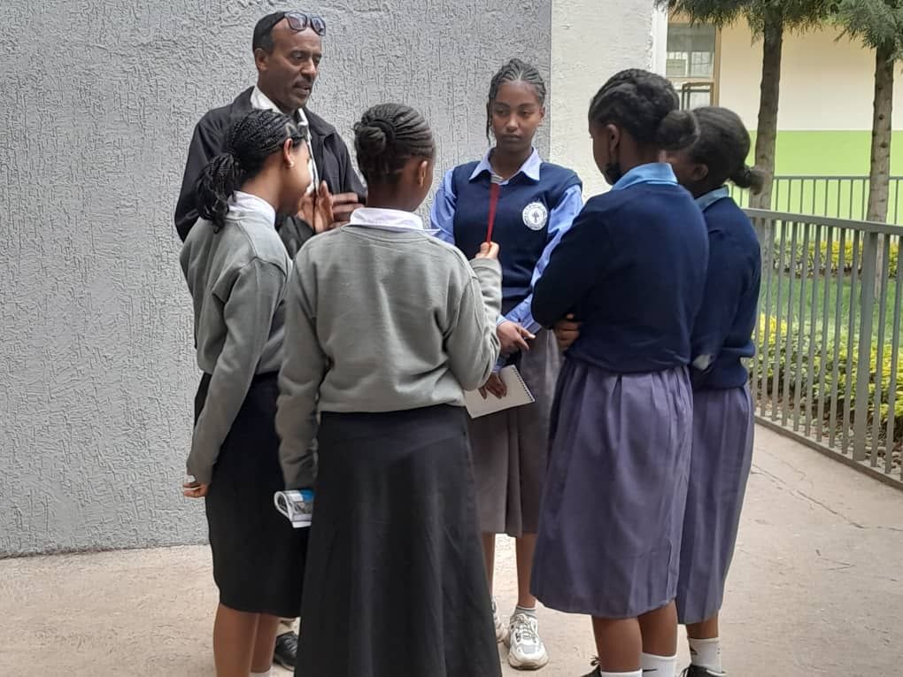 Mentor speaking with five students in school uniforms in an outdoor courtyard.