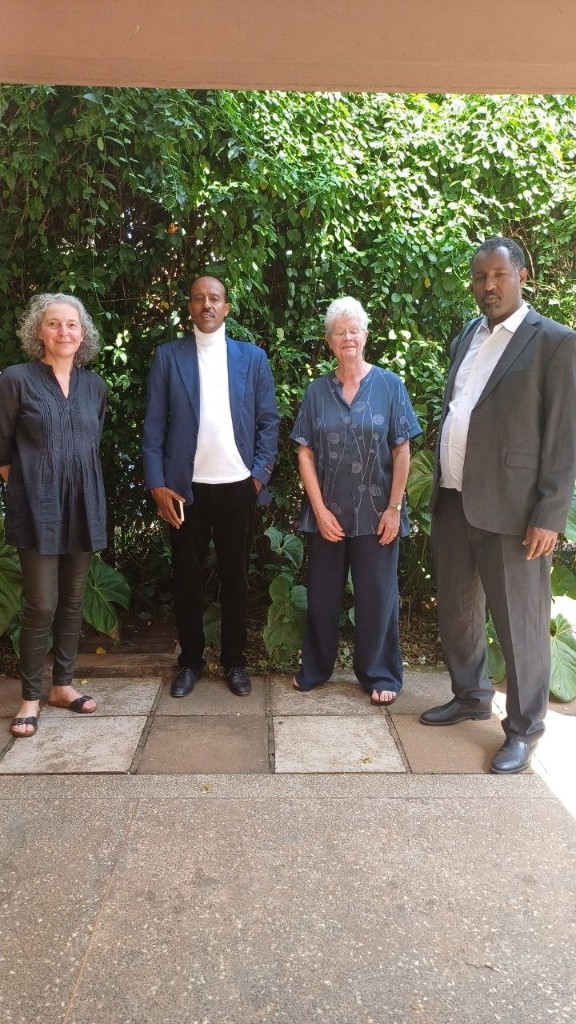 Four people standing outdoors in front of dense green foliage.